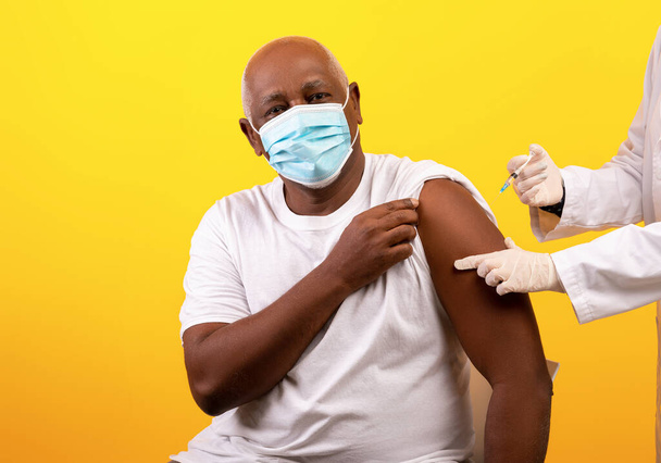 Γιατρός που δίνει εμβόλιο κορωναϊού σε έναν ηλικιωμένο αφροαμερικανό ασθενή σε φόντο πορτοκαλί στούντιο. - Φωτογραφία, εικόνα