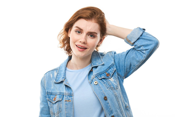 A girl student with red hair happily thinks and looks at the camera while holding her finger near her chin. Pensive thoughtful woman in a blue denim jacket on a white background. Copy space - Fotó, kép