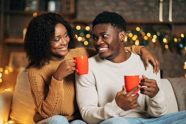 Χαρούμενο μαύρο ζευγάρι αγκαλιάζει και πίνει καφέ, κοιτάζεται, κάθεται στον καναπέ σε διακοσμημένο δωμάτιο τα Χριστούγεννα - Φωτογραφία, εικόνα