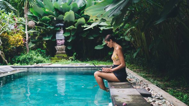 Side view of focused female in summer clothes sitting on poolside with legs in pool while working on laptop in tropical resort hotel in daylight - Fotoğraf, Görsel