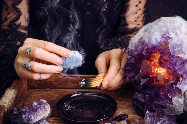 Glamorous woman in black dress cleansing crystals gemstones by smudging Palo Santo wood stick. Remove negative energy. - Photo, Image