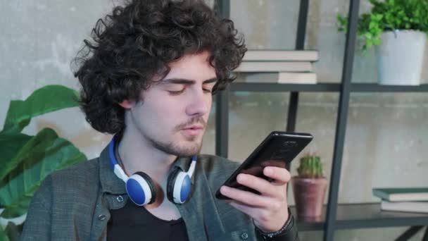 Junge Kaukasier sitzen am Schreibtisch im Home Office und unterhalten sich per Lautsprecher auf dem Smartphone. Millennial-Männchen nutzen Handy, um Sprachnachrichten aufzuzeichnen. - Filmmaterial, Video