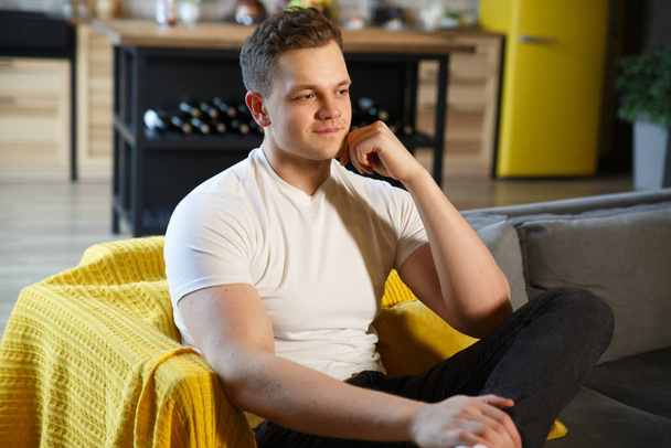 Young attractive man in casual clothes is sitting on the sofa in a cozy home interior. - Фото, зображення