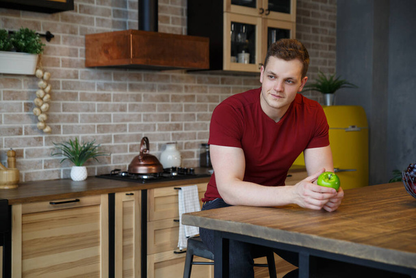 Young attractive guy with an apple in his hand in the interior of the kitchen. - Foto, Bild
