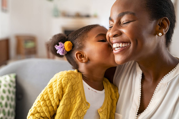 Smiling mature african american mother helping daughter sitting on lap putting money in piggy bank. Cute little black girl saving money by adding a coin in piggy bank with mother at home. Daughter sitting on moms laps and putting coins into piggybank - Zdjęcie, obraz