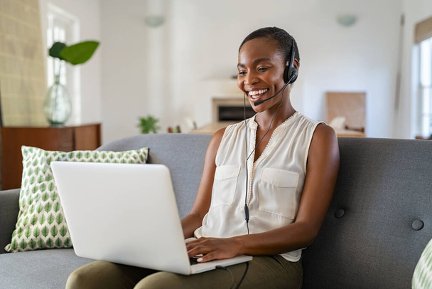 Successful middle aged african american woman with headphone and mic talking on video call using laptop. Cheerful mid black woman enjoy video call on laptop while sitting on couch at home. Happy smiling lady using computer for video conference while  - Fotó, kép