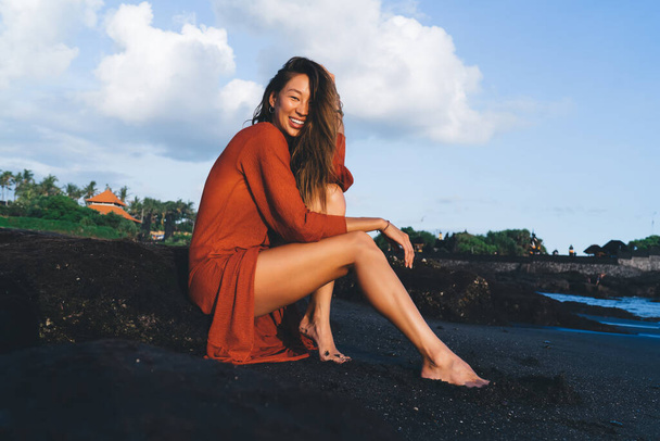 Low angle of young cheerful Asian female tourist in light dress looking at camera and smiling while recreating on sandy beach near ocean in Bali - Valokuva, kuva