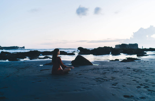Young Asian woman sitting in meditation pose on sandy shore with rocks in ocean and looking away during morning hour - Foto, afbeelding