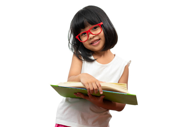 Happy Asian little preschool girl wearing red glasses holding and read a green book on white isolated background. Concept of school kid and education in elementary and preschool, home school - Photo, Image