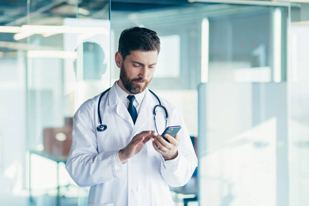 Male doctor in a white coat in a modern clinic reads information from a mobile phone corresponds with colleagues happy and smiles - Zdjęcie, obraz