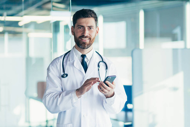 Male doctor in a white coat in a modern clinic reads information from a mobile phone corresponds with colleagues happy and smiles - Zdjęcie, obraz