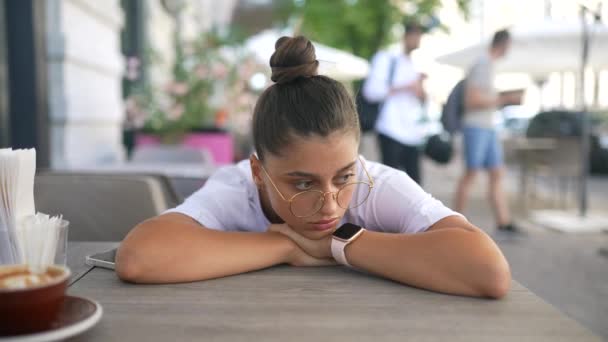 Znudzona, obojętna dziewczyna przy stole w ulicznej kawiarni - Materiał filmowy, wideo