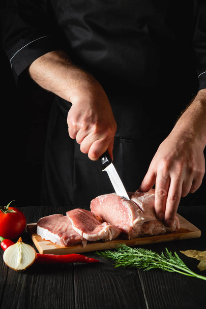 Šéf řeznictví krájí vepřové maso nožem v kuchyni, vaří jídlo. Zelenina a koření na kuchyňském stole připravit lahodný oběd - Fotografie, Obrázek