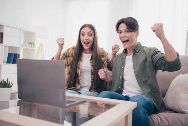 Photo of lucky attractive young couple wear casual outfits smiling rising fist reading modern gadget sitting couch indoors house room. - Фото, зображення