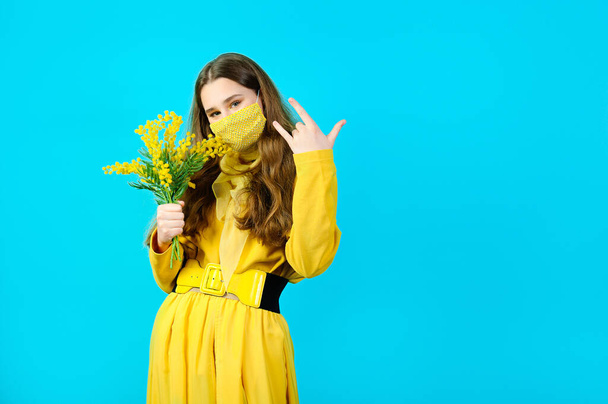portrait of a teenage girl in a glamorous mask on her face with mimosa flowers in her hands. blue background - Photo, Image