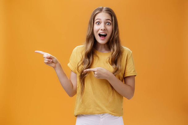 Portrait of excited surprised and amazed cute young european slim woman with wavy natural hairstyle dropping jaw astonished pointing left asking question about awesome thing over orange wall - Photo, Image