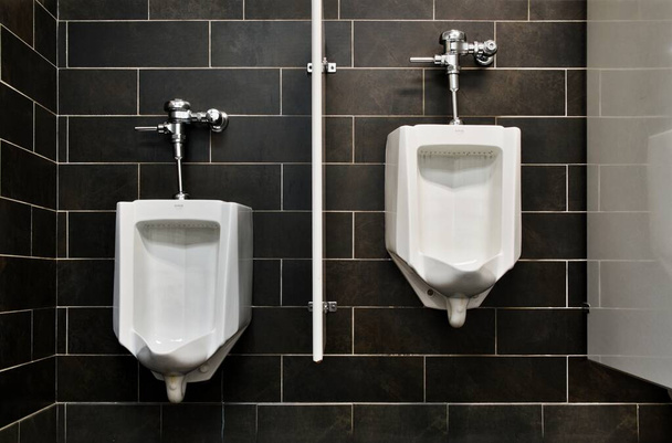 Zwei leere Pissoirs in einer öffentlichen Toilette mit schwarzem Fliesenhintergrund. - Foto, Bild