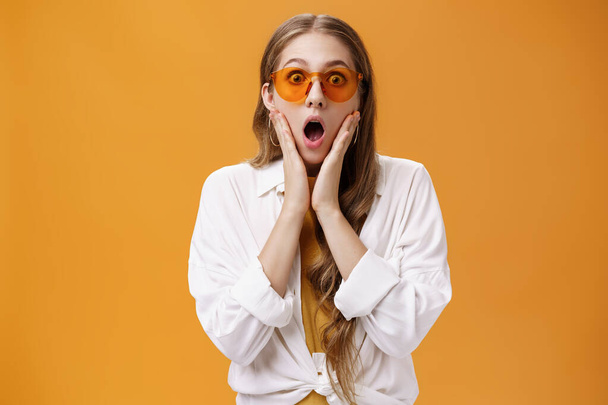 Waist-up shot of amazed and surprised party girl in trendy sunglasses and cool outfit gasping from shock standing with opened mouth concerned look and hands on face posing over orange background - Valokuva, kuva