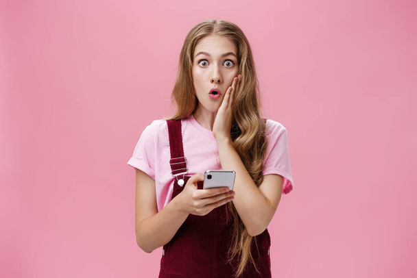 Талия снимок удивлённой ошеломленной юной женщины с волнистыми волосами в комбинезоне, касающейся щеки от шока, держащей смартфон, реагирующий на впечатляющие неожиданные новости через розовую стену - Фото, изображение