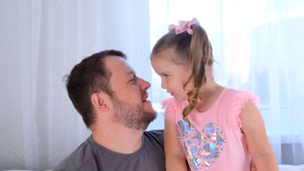 little cute daughter, 3 years old, tells her dad a secret, play together at home. Fathers day. Handsome young man at home with his little cute girl are having fun together. Fatherhood concept, happy - Photo, Image