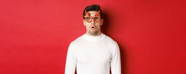 Primer plano del hombre sorprendido e impresionado en suéter blanco, gafas de fiesta, diciendo wow y mirando asombrado a la cámara, de pie sobre el fondo rojo - Foto, imagen