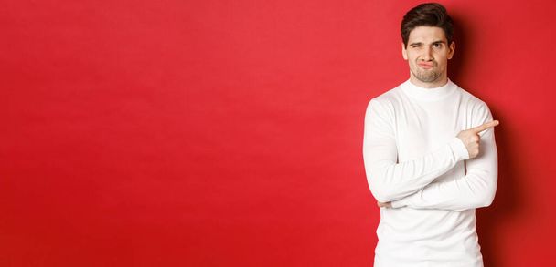 Doubtful and reluctant handsome man grimacing, pointing fingers right at something bad, standing in white sweater over red background - Foto, immagini