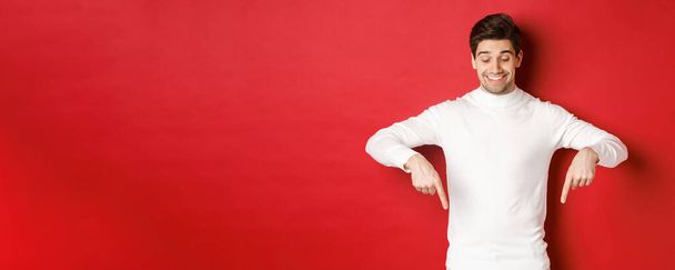 Immagine di uomo di bell'aspetto sognante in maglione bianco, guardando e puntando il dito verso il basso lo spazio copia, pubblicità sul nuovo anno e le vacanze, in piedi su sfondo rosso - Foto, immagini
