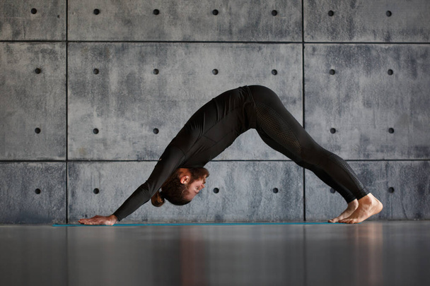 Nuori parrakas mies mustissa urheiluvaatteissa harjoittelee joogaa harmaata seinää vasten.. - Valokuva, kuva