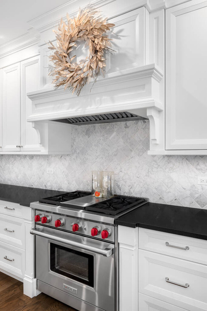 CHICAGO, IL, USA - OCTOBER 12, 2020: A luxurious white kitchen detail with a stainless steel Wolf stove, black granite counter tops, and a wreath hanging from the white oven hood. - Photo, Image