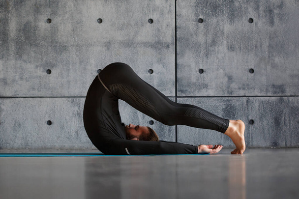 A young bearded man in black comfortable sportswear is doing yoga indoors. - Foto, afbeelding