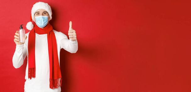 Концепция ковида-19, Рождества и праздников во время пандемии. Счастливый и довольный мужчина в шляпе Санты и медицинской маске, показывающий большой палец вверх в одобрении и рекомендующий дезинфицирующее средство для рук - Фото, изображение