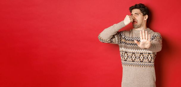 Imagen de hombre guapo disgustado en suéter de invierno, rechazando algo con mal olor, cerrar la nariz y levantar la mano para declinar, de pie sobre el fondo rojo - Foto, imagen