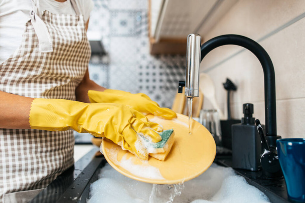Young adult woman with yellow protective gloves washing her dishes on kitchen sink. Household and home hygiene routine. - Foto, Imagen