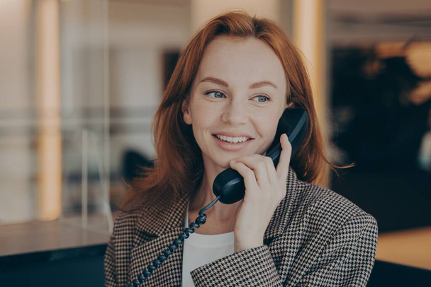 Portrait d'une travailleuse de bureau satisfaite utilisant un téléphone fixe noir, appelant un partenaire d'affaires - Photo, image