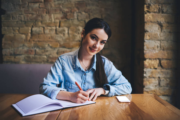 Smiling dark haired woman in jean shirt writing in notebook and looking at camera in modern cafe with brick wall on background - Foto, immagini