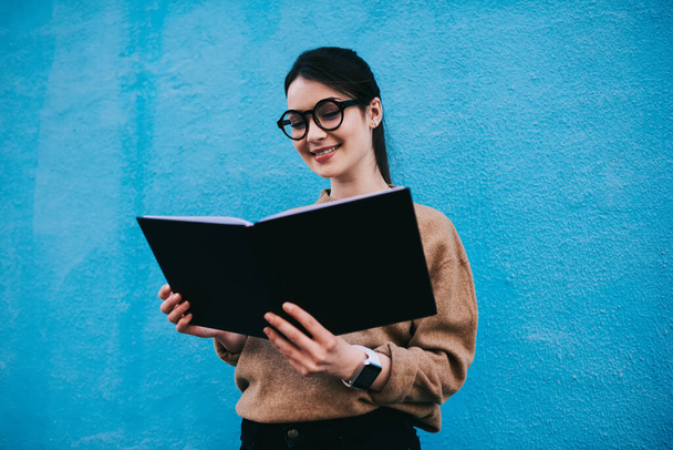 Positive female in casual outfit with toothy smile and eyewear standing near blue wall with opened book and reading with interest - Foto, imagen
