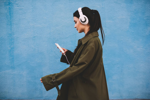 携帯電話を閲覧しながらストリートを歩き、ヘッドフォンで音楽を聴く流行の服で穏やかな女性の側のビュー - 写真・画像