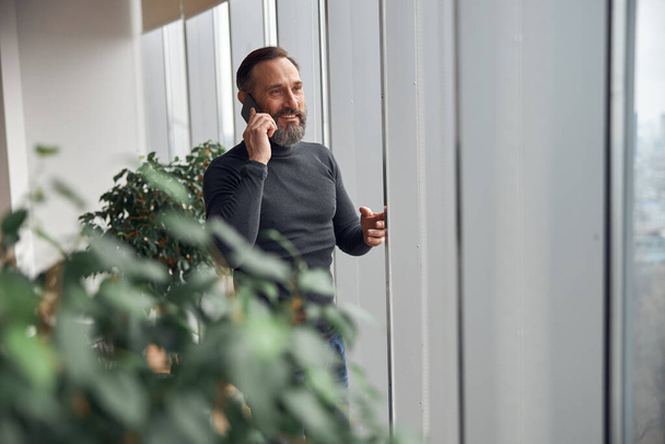 Счастливый умный взрослый кавказский бизнесмен стоит возле больших окон в офисном пространстве и держит смартфон - Фото, изображение