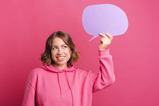 Young trendy woman in pink hoodie thinking and holding blank violet speech bubble - Foto, Imagen