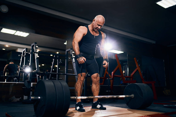 Older powerlifter preparing to exercise deadlift with barbell while on cross training in a gym. - Photo, image