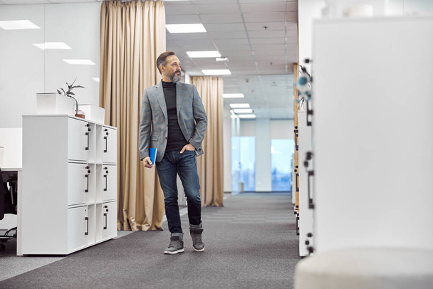 Έξυπνο, χαρούμενο αρσενικό εργάτη περπατά σε ένα σύγχρονο γραφείο. - Φωτογραφία, εικόνα