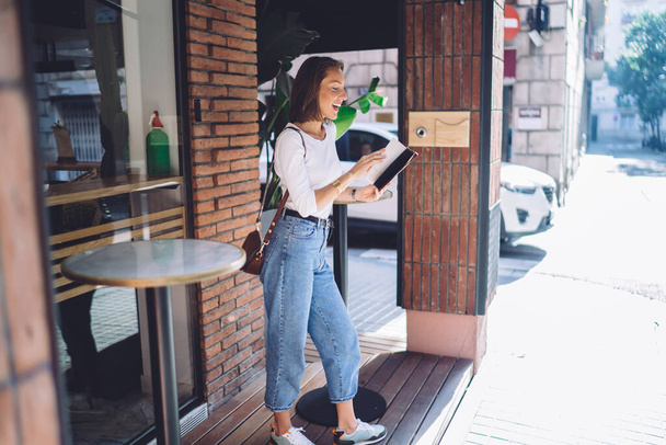 Cheerful caucasian female in casual wear standing on cafe terrace holding new interesting novel for reading, smiling woman 20s enjoying free tin on urban settings checking to do list in diary - Photo, Image