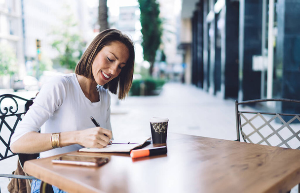 Smiling millennial female in white shirt writing down ideas into planner while sitting at table of summer cafe with cup of drink to go - Fotoğraf, Görsel