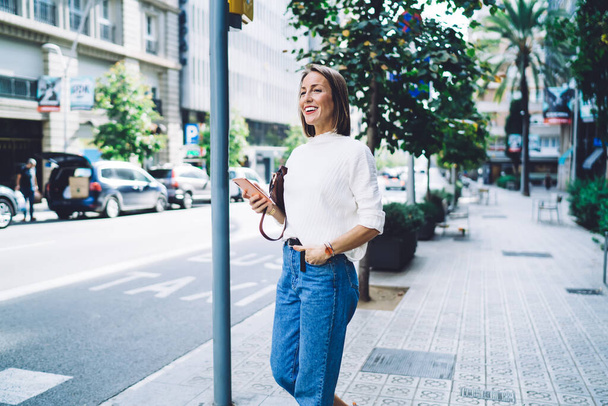 Mooie vrouw in casual outfit met behulp van mobiele telefoon staan met de hand in zak op de stoep klaar om over te steken straat weg te kijken en glimlachen - Foto, afbeelding