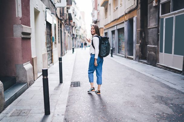 Plná délka portrét veselá žena kráčí na ulici města a usmívá se na kameru, šťastná žena turista s smartphone gadget pomocí roaming internetu pro vyhledávání umístění čtvrti ve městě - Fotografie, Obrázek