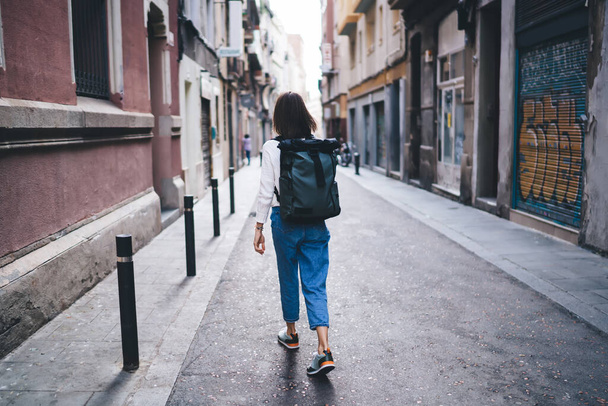 Back view of woman in casual wear in sneakers with backpack on back and walking along street of residential area - Φωτογραφία, εικόνα