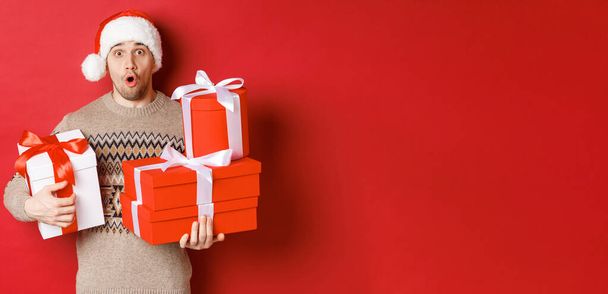 Concept of winter holidays, new year and celebration. Image of surprised handsome guy in sweater and santa hat, receiving gifts, holding boxes with presents and looking amazed - Photo, Image