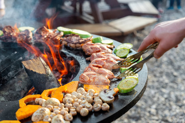 Close-up detail view of chef flipping by tongs tasty crispy marinated quails and vegetables grilled at round steel iron firepit hearth table surface brazier with burning firewoods. Barbecue yard home - Photo, Image