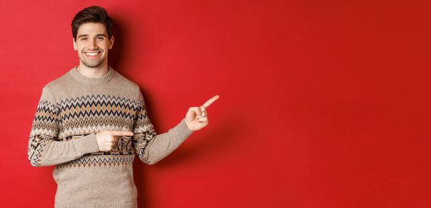 Immagine di attraente uomo sorridente, indossa maglione di Natale, indicando le dita a destra e mostrando pubblicità di nuovo anno, in piedi su sfondo rosso - Foto, immagini