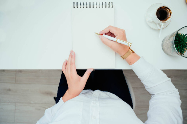 Female hands with a notebook and pen, coffee and plant on white desk table, top view. Woman writing, taking notes. Top view with copy space - Foto, afbeelding